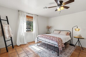 Ein Schlafzimmer mit einem Bett, einem Deckenventilator, einem Fenster mit Vorhängen, einem Nachttisch und einer Stehlampe.