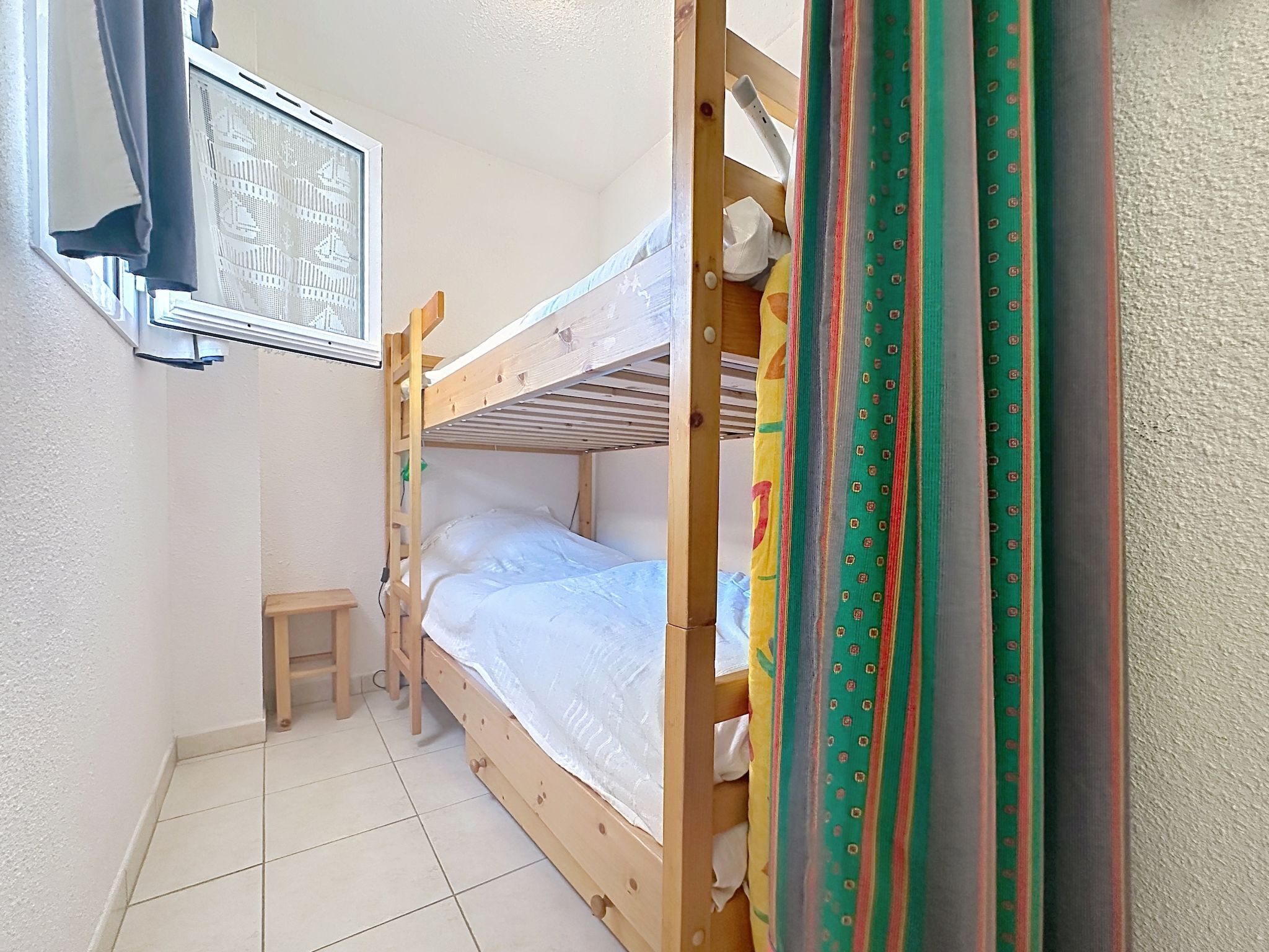 A small, narrow room with a bunk bed, a small stool, and a window with a view of a building.