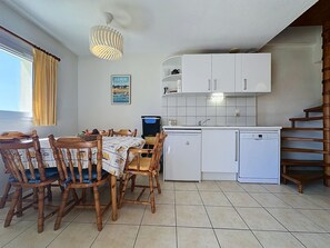 A compact kitchen with white cabinets, a small refrigerator, and a dining area with wooden chairs and a table. - Port Saint-Jacques by Interhome (La Bernerie-en Retz)