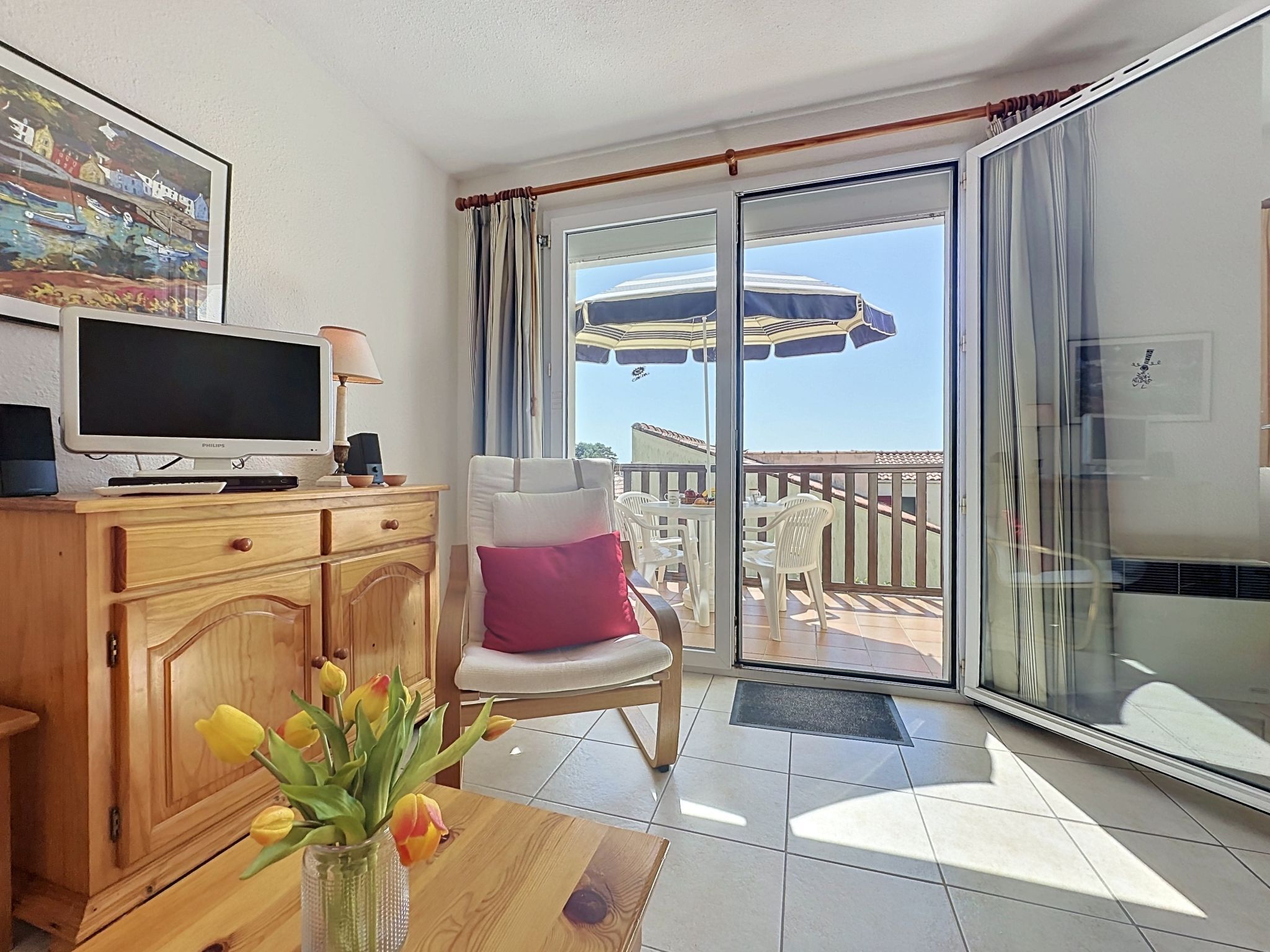 A room with a television, a wooden cabinet, a chair with a pink cushion, a vase with flowers, and a view of an outdoor patio with umbrellas.