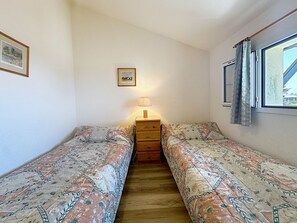 Una habitación pequeña y estrecha con dos camas individuales, un aparador de madera, una lámpara de pared y un ventanal con cortinas.