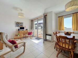 A living room with a TV, seating, a dining table, and a view through a glass door. - Port Saint-Jacques by Interhome (La Bernerie-en Retz)