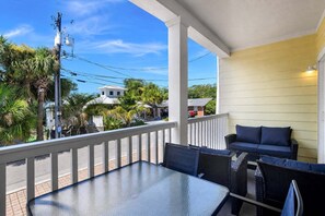 A covered outdoor seating area with a table and chairs, overlooking a street with palm trees and buildings. - Bright Coastal Retreat on Tybee's South End | Heated Pool Access (Tybee Island)