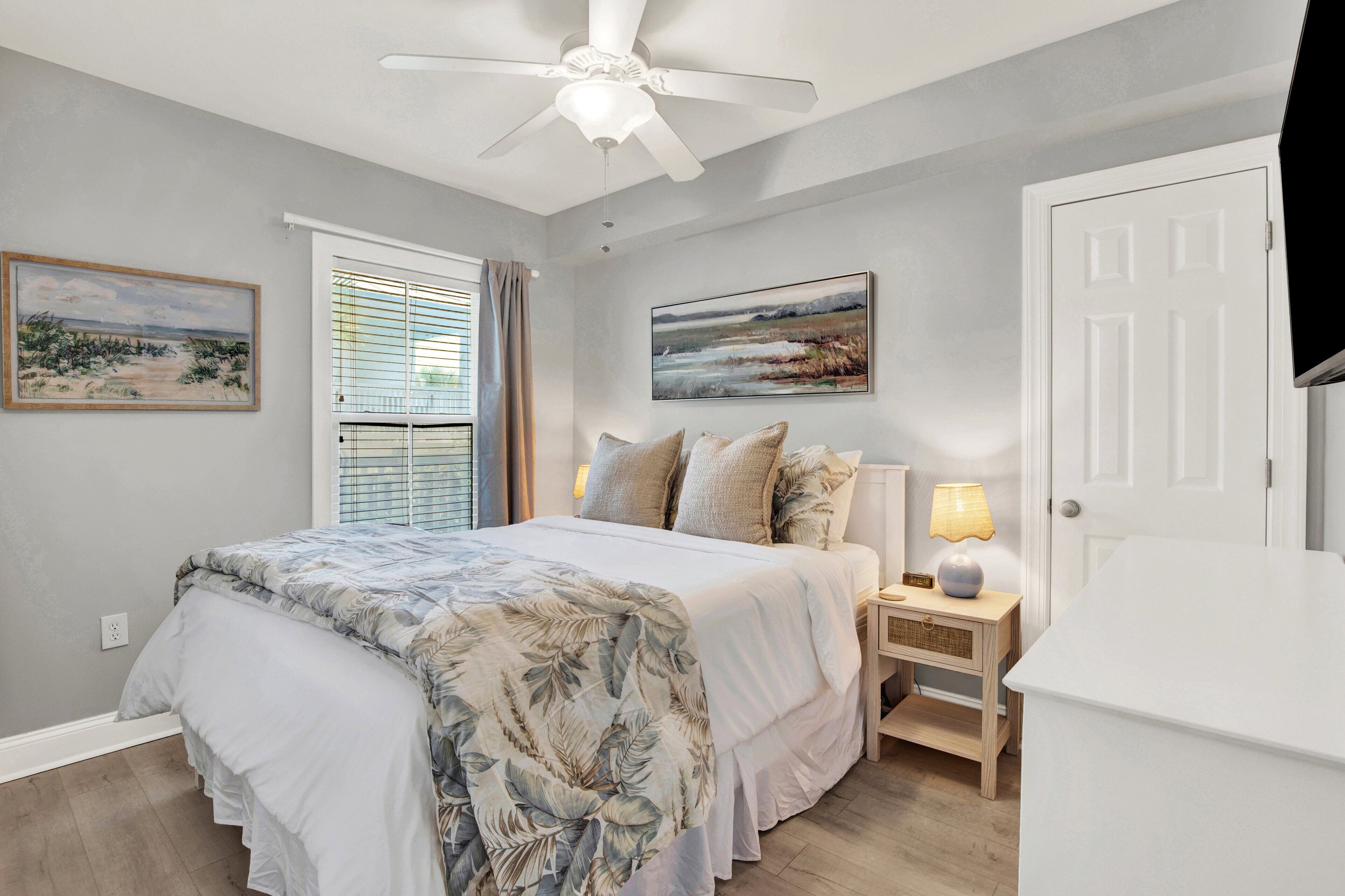 A bedroom with a bed, a nightstand, a lamp, a ceiling fan, and two paintings on the wall.
