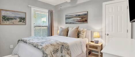 A bedroom with a bed, a nightstand, a lamp, a ceiling fan, and two paintings on the wall.
