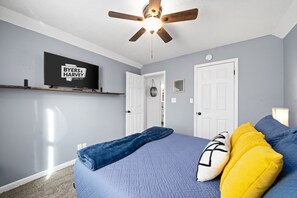 Quarto com cama, ventilador de teto, televisão montada e uma porta.