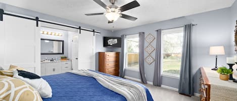 A bedroom with a bed, a ceiling fan, a TV, a dresser, a window with curtains, and a view of the outdoors.