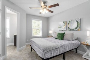 A bedroom with a bed, a ceiling fan, a window, and two abstract paintings on the wall.