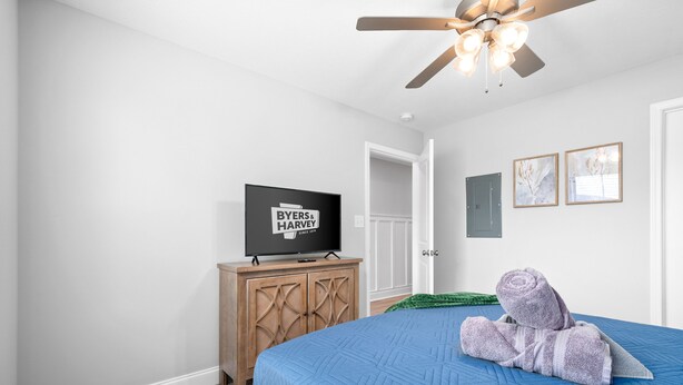 Una camera da letto con una coperta azzurra trapuntata, un mobiletto in legno con un televisore e una ventola a soffitto.
