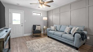Un soggiorno con un divano grigio, una sedia di legno, una ventola a soffitto e un tappeto decorato.