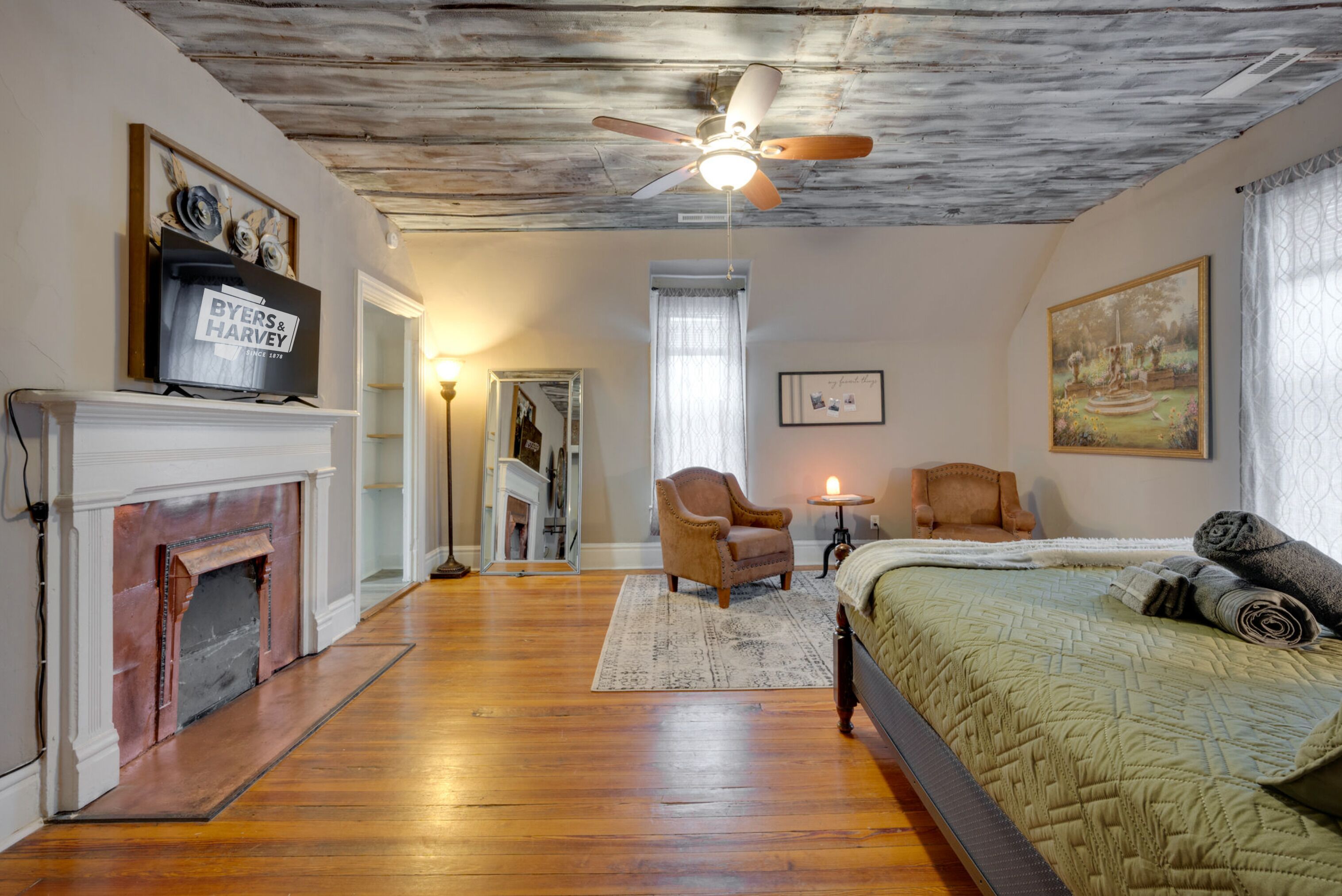A room with a fireplace, a bed, a ceiling fan, and a painting on the wall.