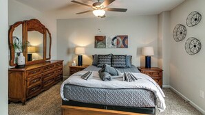Quarto com cama, cômoda de madeira, duas mesas de cabeceira com abajures, ventilador de teto e quadros nas paredes.