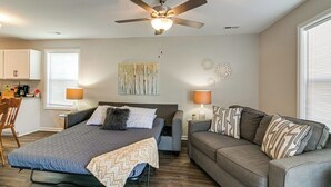Une chambre à coucher moderne avec un lit, un canapé gris, un ventilateur de plafond et un coin cuisine à l’arrière-plan.