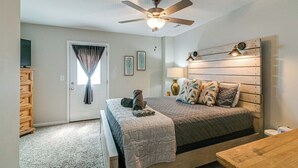 Une chambre à coucher comprenant un lit, un ventilateur de plafond, une fenêtre avec des rideaux, une commode en bois et une table de chevet.