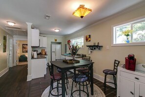 A compact kitchen with white cabinets, a dining table with chairs, and a window with a vase of flowers.