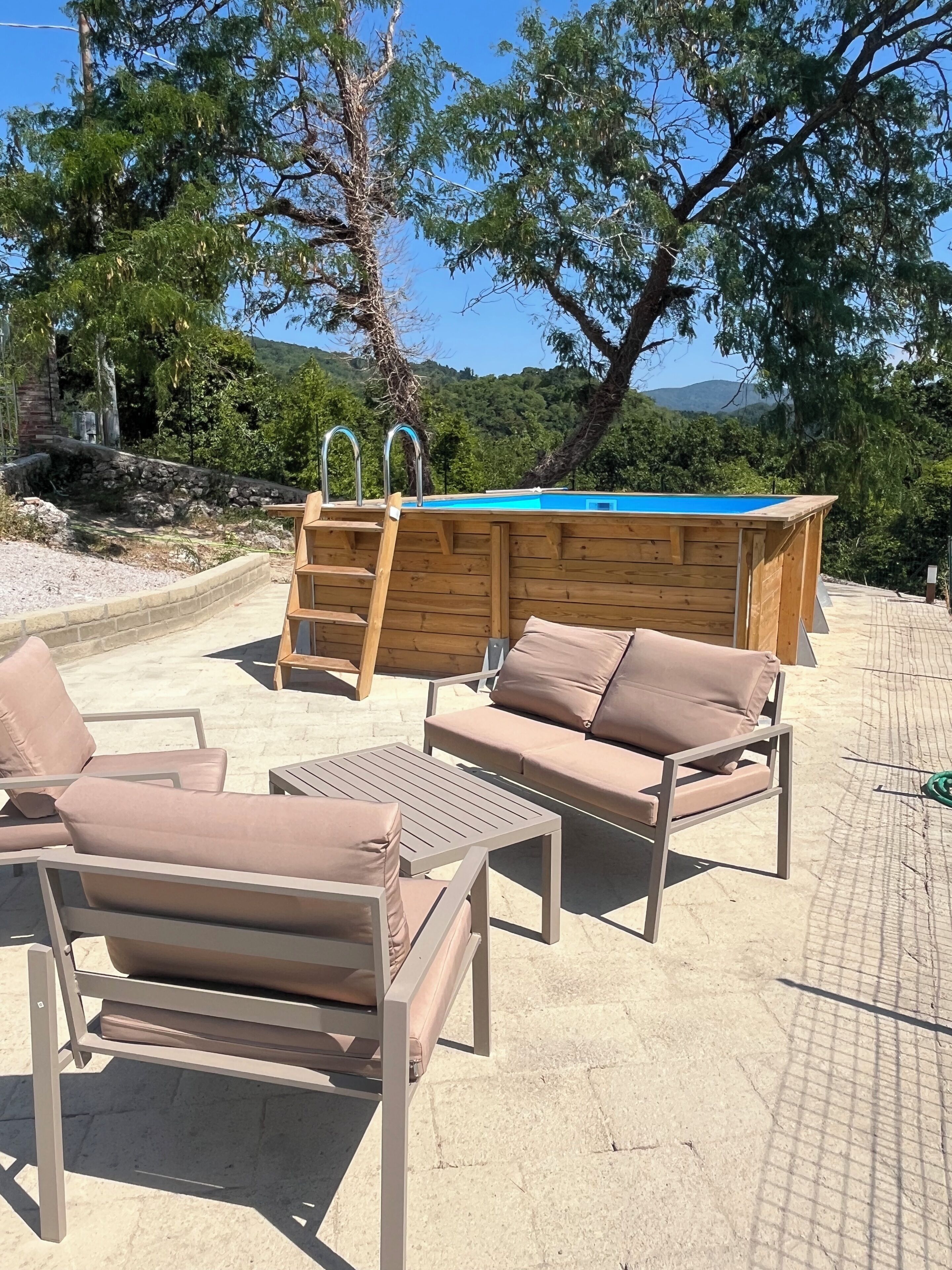 An outdoor pool area with wooden loungers and a ladder, surrounded by trees and a mountainous backdrop.