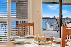 A dining area with a table set for four, a wooden chair, and a view of the sea through a large window. - Waterfront Games House | Pool & Bay Views (Koolewong)