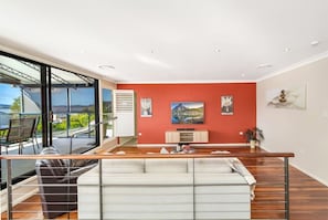 A modern living room with a large window, a sofa, a television, and a fireplace. - Waterfront Games House | Pool & Bay Views (Koolewong)
