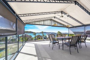 A covered outdoor dining area with a view of the sea and hills. - Waterfront Games House | Pool & Bay Views (Koolewong)