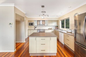 A modern kitchen with a central island, stainless steel appliances, and a window providing natural light. - Waterfront Games House | Pool & Bay Views (Koolewong)