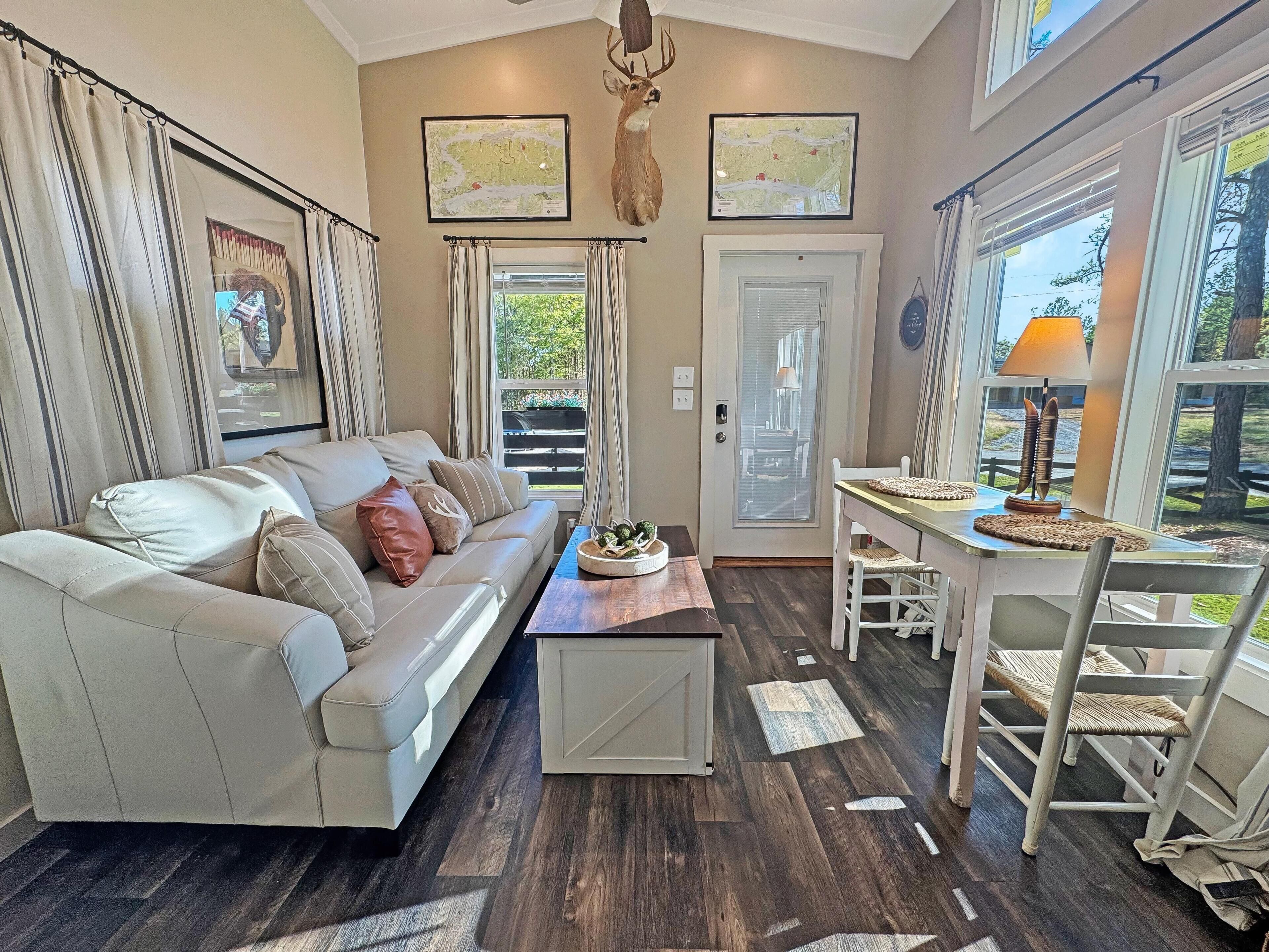 A living room with a sofa, a coffee table, a dining table with chairs, a deer head decoration, and two framed maps on the wall.