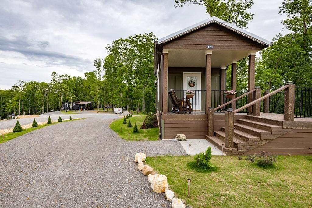 A wooden cabin with a porch and steps leading to a door, surrounded by a gravel driveway and small trees.