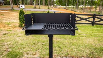 A black barbecue grill with a slotted grate, set on a concrete base, outdoors with a grassy area and trees in the background.
