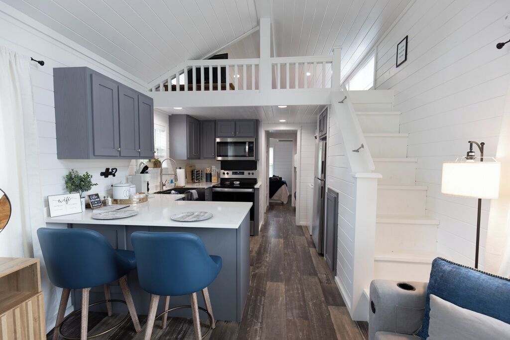 Une cuisine moderne avec des meubles gris, un îlot central doté de chaises bleues et un escalier menant à un niveau supérieur.