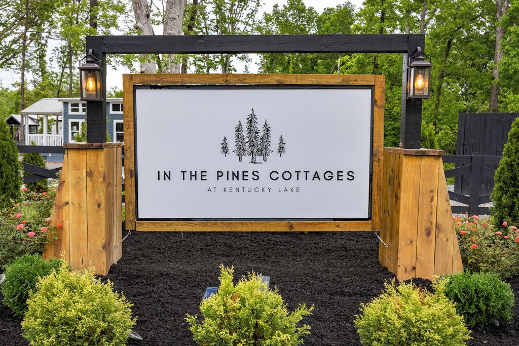 Placa para 'In the Pines Cottages' no Lago Kentucky, rodeada por canteiros de jardim ajardinados e estruturas de madeira.