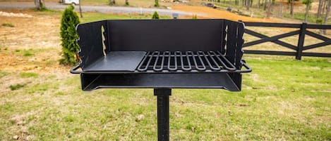 A black barbecue grill with a slotted grate, set on a concrete base, outdoors with a grassy area and trees in the background.