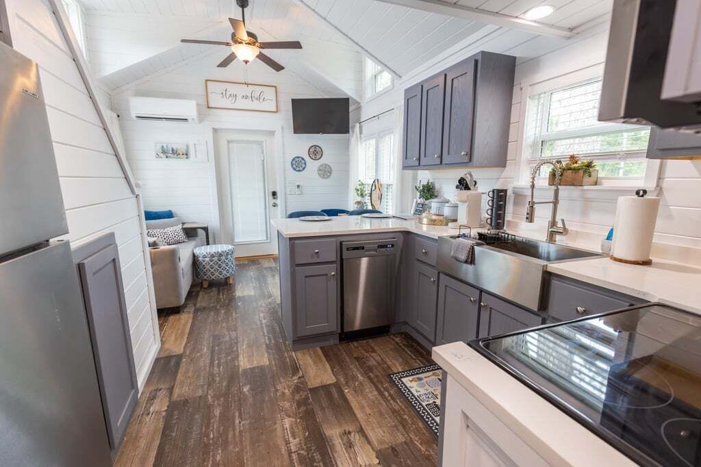 A kitchen with grey cabinets, a dishwasher, and a stove. There is a ceiling fan and a window with plants on the counter.