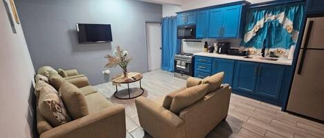 Sala de estar com sofá e uma pequena mesa redonda, cozinha com armários e eletrodomésticos azuis, e um ventilador de teto.