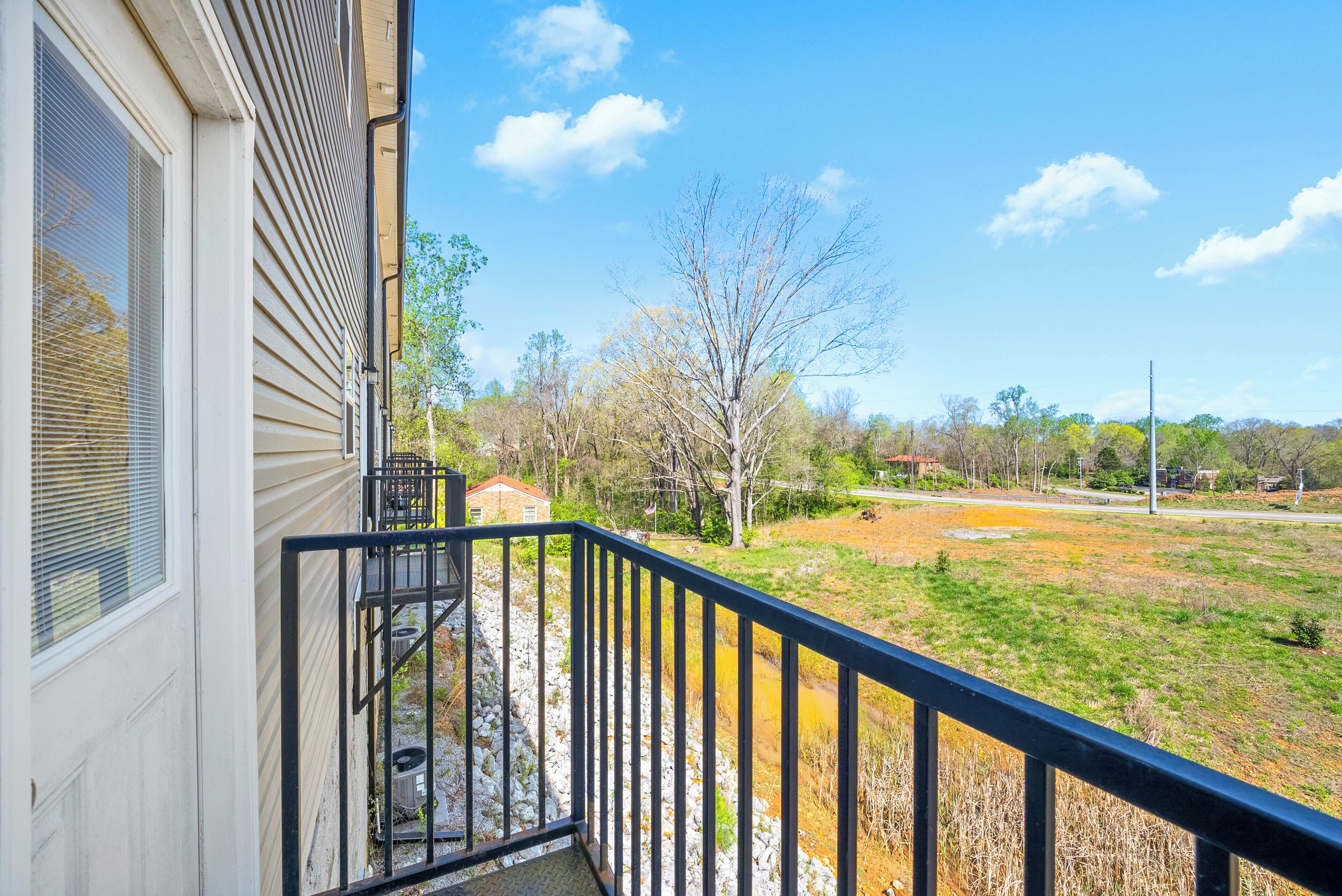 A balcony with a metal railing overlooking a grassy area with trees and a distant road.