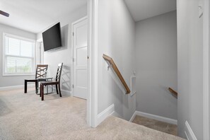 Ein Zimmer mit an der Wand montiertem Fernseher, einem Esstisch mit Stühlen, einer Treppe mit Holzhandläufen und einem Fenster mit Jalousien.