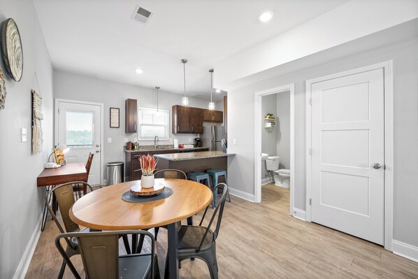 A modern kitchen with wooden cabinets, a dining area with a round table, and a view into the adjacent bathroom.