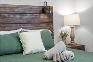 A bed with a wooden headboard, green bedding, a rolled towel, and a nightstand with a lamp and vase. - Birdie's Nest on Golf Club (1625 Golf Club Ln) (Clarksville)