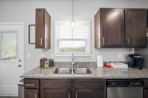 A kitchen with a granite countertop, stainless steel sink, dark wooden cabinets, a window with blinds, and a coffee maker. - Birdie's Nest on Golf Club (1625 Golf Club Ln) (Clarksville)
