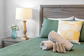 A neatly made bed with a green bedspread, a rolled towel, and a pillow with a yellow and white pattern.