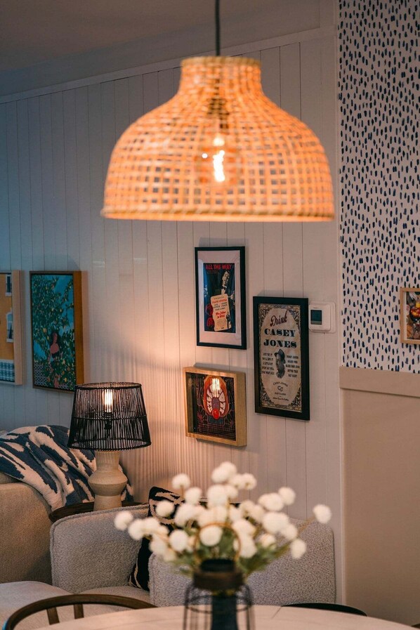 Une pièce chaleureuse avec une table ronde, un vase rempli de fleurs blanches, une lampe suspendue et des tableaux encadrés accrochés au mur.