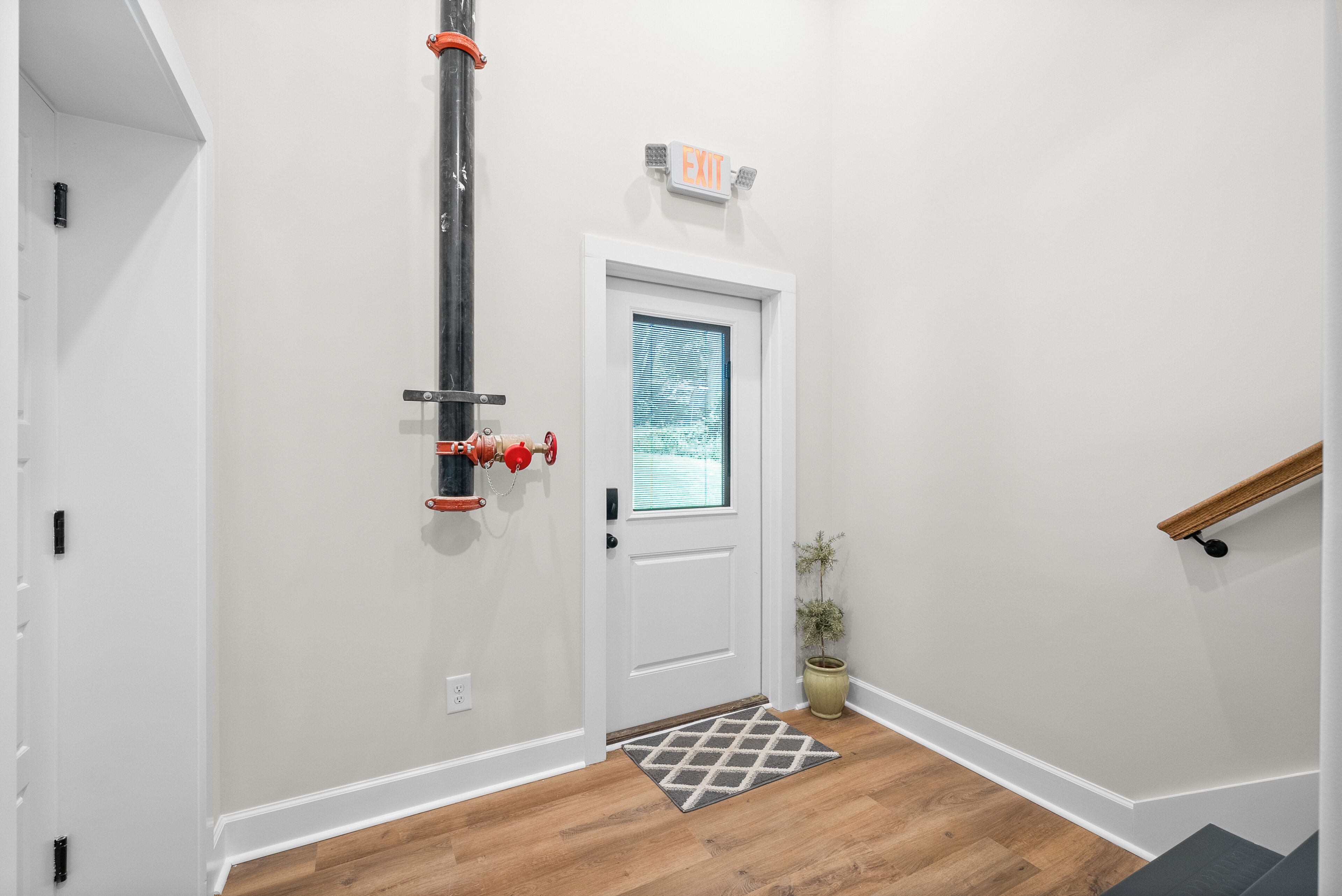A hallway with a white door, a small rug, a potted plant, and an exit sign.