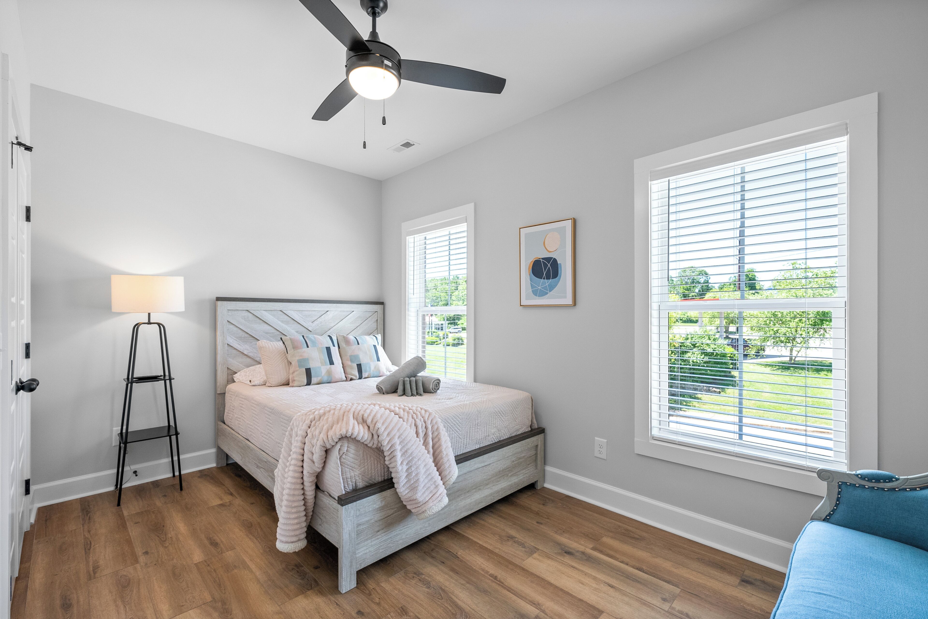 Un dormitorio con cama, lámpara de pie, ventilador de techo y dos ventanas con persianas.