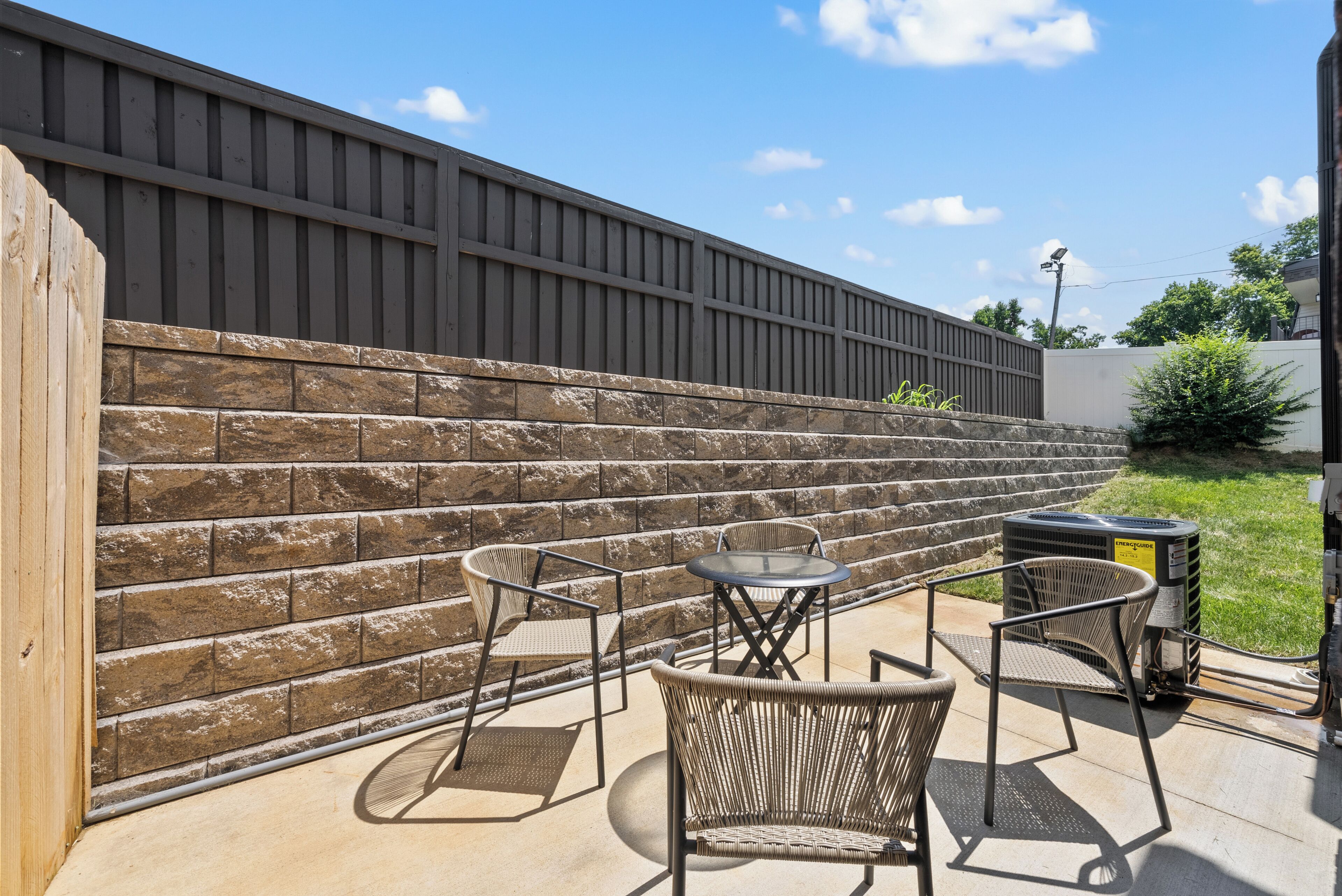 A patio area with a stone wall, two wicker chairs, a small round table, and an air conditioning unit.