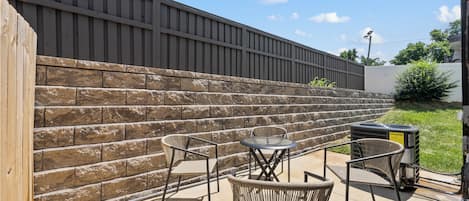 A patio area with a stone wall, two wicker chairs, a small round table, and an air conditioning unit.