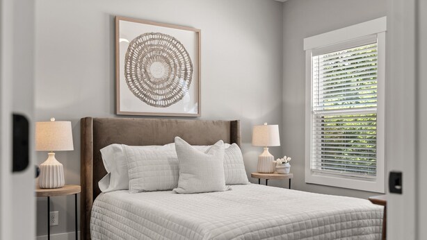 A bedroom with a large bed, two bedside tables with lamps, a framed artwork above the bed, and a window with blinds. - River's Edge (111 Hickory Grove BLVD) (Clarksville)