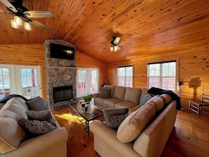 A log cabin interior with a stone fireplace, a ceiling fan, and a large window. - Cottage on Cypress Bay near Paris Landing State Park (Buchanan)