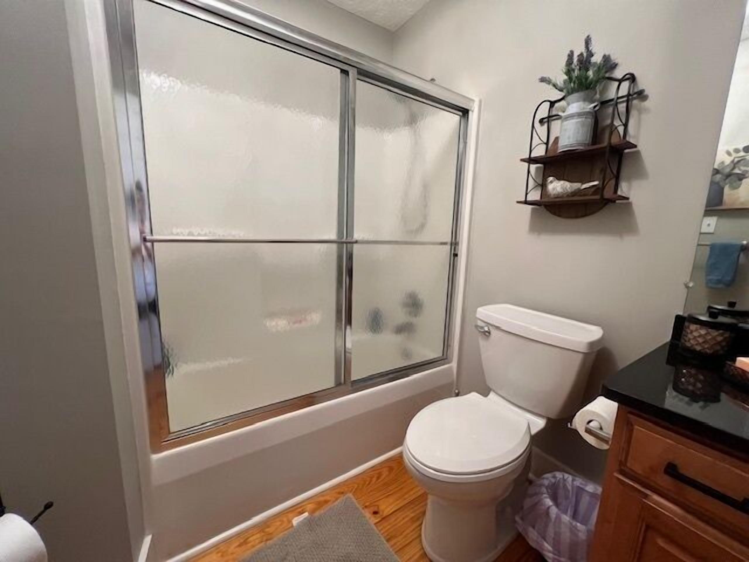 A bathroom with a toilet, a window with frosted glass, a wooden floor, and a shelf with a potted plant.