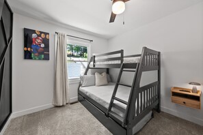 A bedroom with a bunk bed, a window with curtains, a ceiling fan, and a wall-mounted shelf with a lamp.