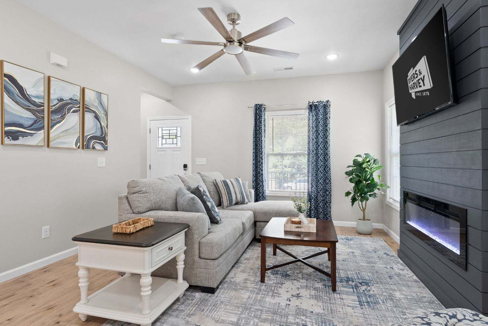 A living room with a grey sofa, a white coffee table, a wooden side table, a ceiling fan, a fireplace, and a large window with blue curtains.