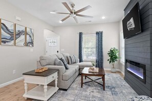 A living room with a grey sofa, a white coffee table, a wooden side table, a ceiling fan, a fireplace, and a large window with blue curtains.