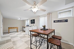 Ein Zimmer mit kariertem Boden, einem Deckenventilator, einem Tisch mit Stühlen, einer Waschmaschine und einem Schild mit der Aufschrift 'WELCOME' an der Wand.